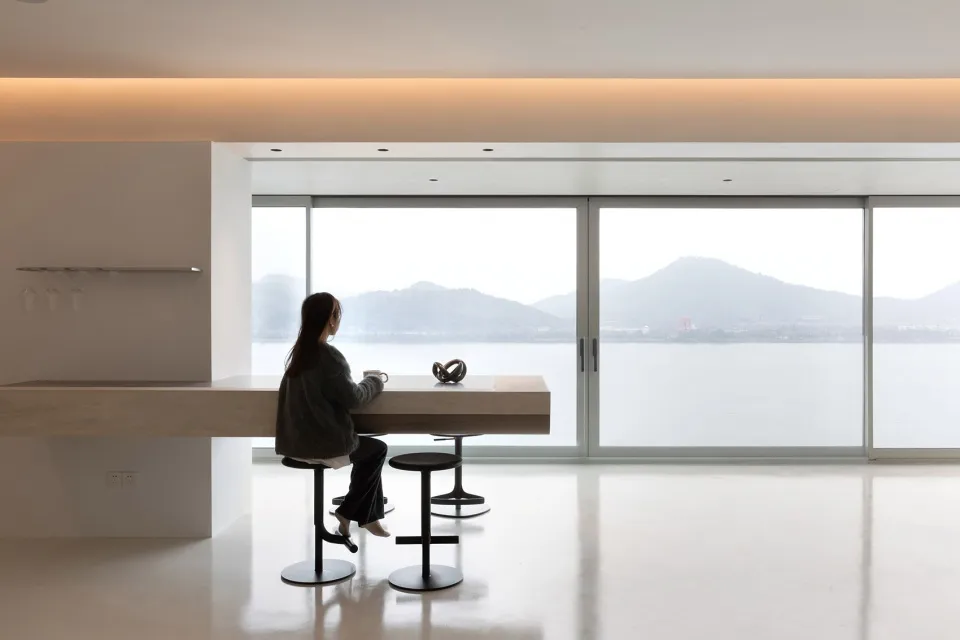 Person sitting at a wooden dining table by a large window overlooking the lake and mountains.