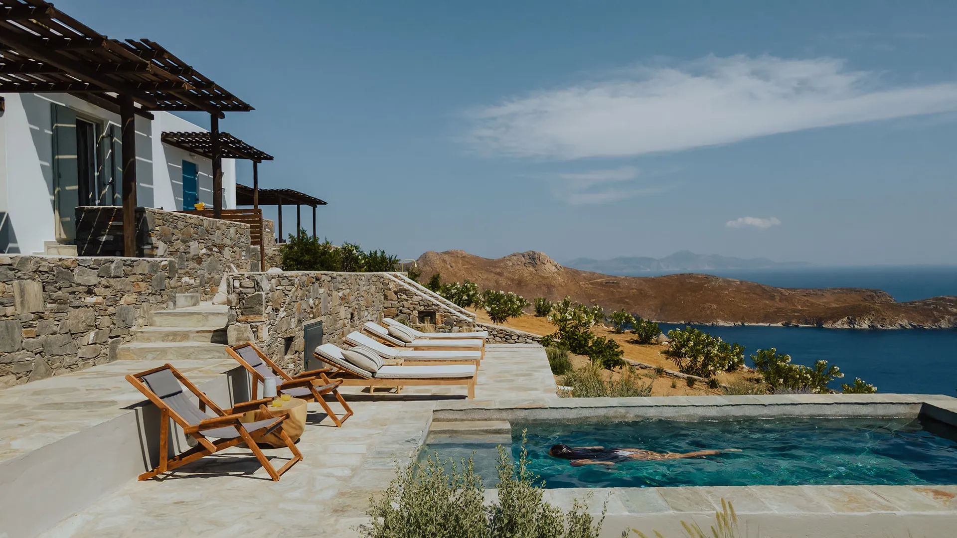 Stone and wooden pool deck with concrete stairs, glass railing, and timber lounge chairs overlooking the ocean.