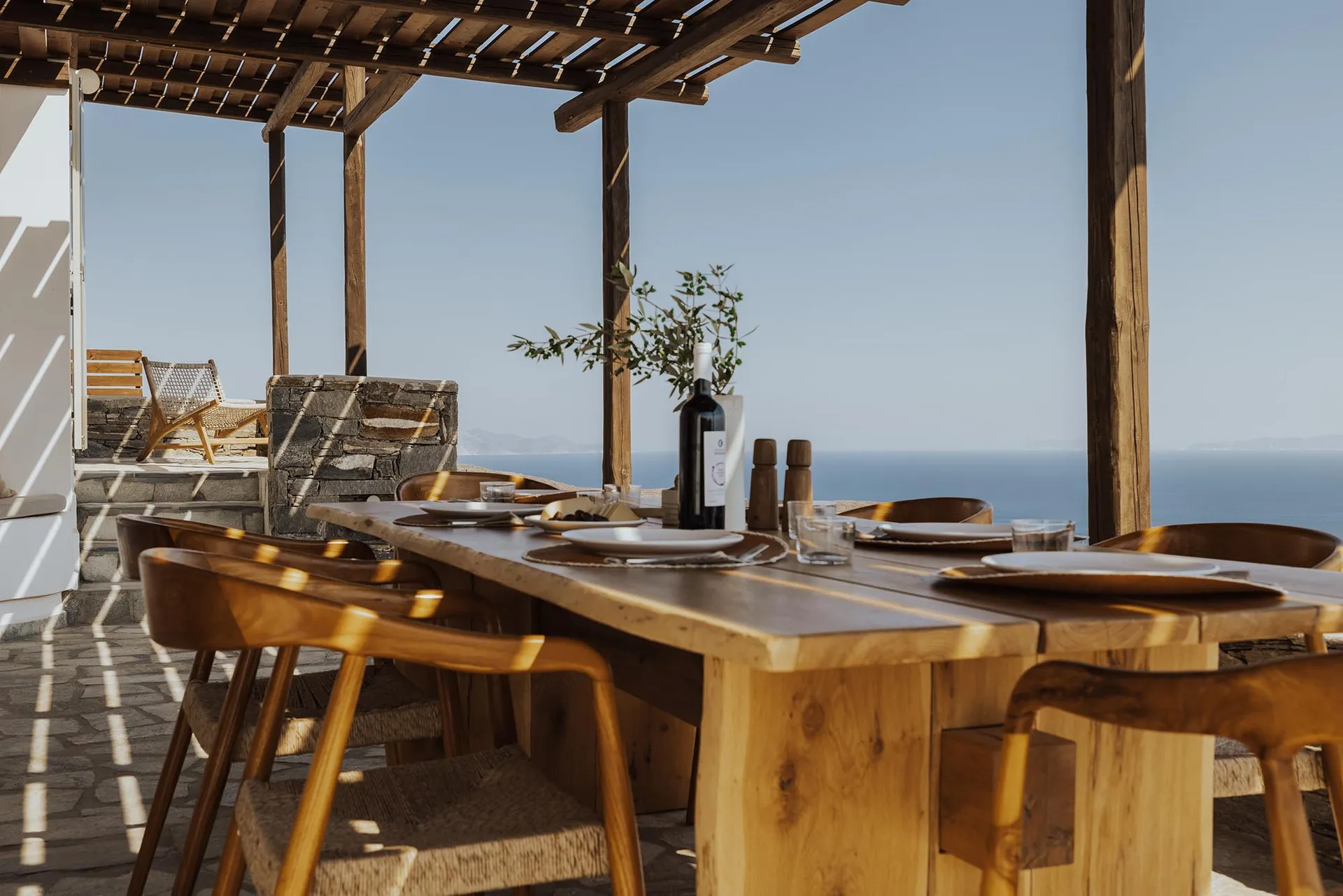A wooden outdoor dining table set with plates, glasses, and a bottle of wine, overlooking the sea.