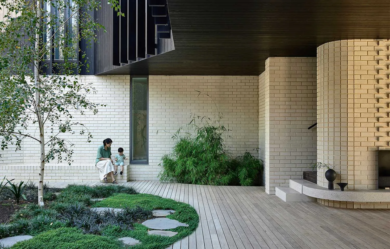 A modern house exterior with a combination of concrete walls and stone elements, surrounded by greenery and a wooden deck.