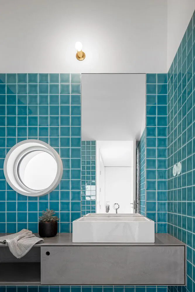 A modern bathroom featuring teal ceramic tiles, a circular window, and a white floating vanity.