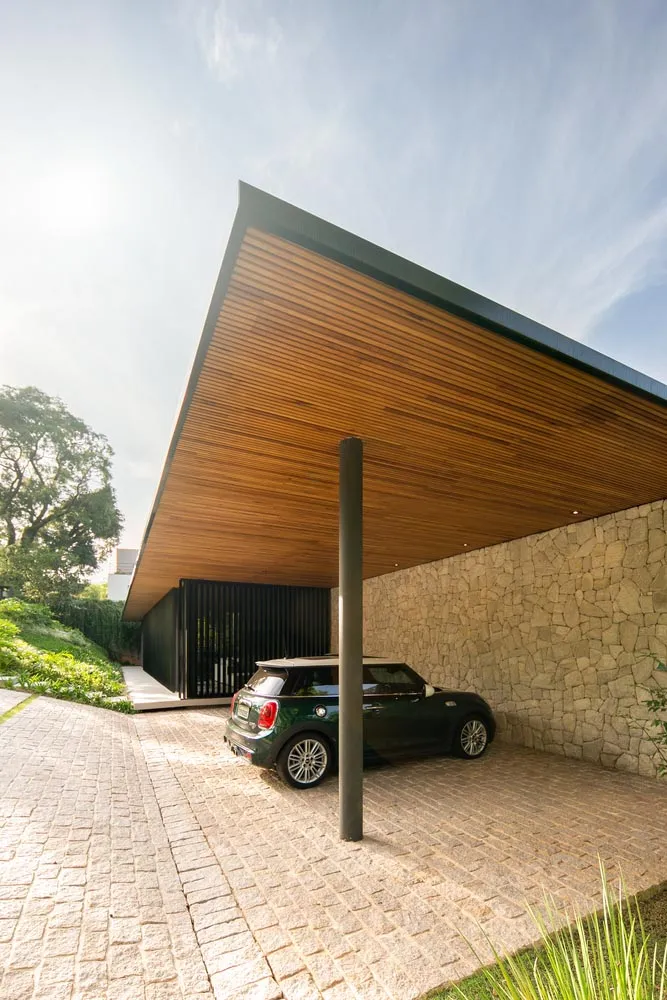 A modern carport with a wooden roof and stone walls featuring a green car parked underneath it on a sunny day.
