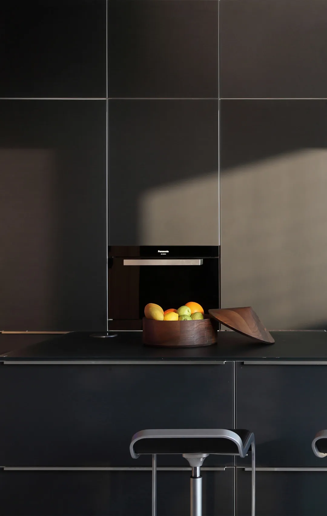 A modern kitchen interior featuring sleek, black cabinets and a countertop, with a wooden bowl containing a variety of fruits