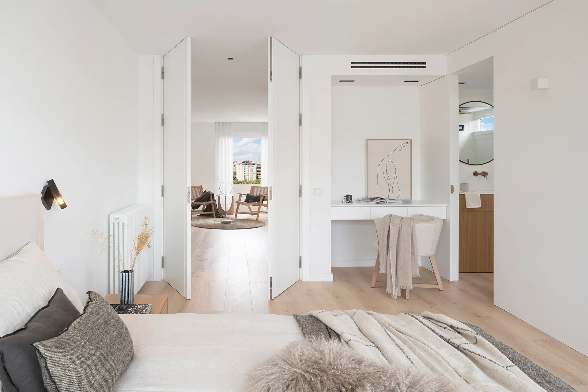 A minimalist bedroom with wooden floors and a bed dressed in neutral tones, featuring a study area and an open layout.