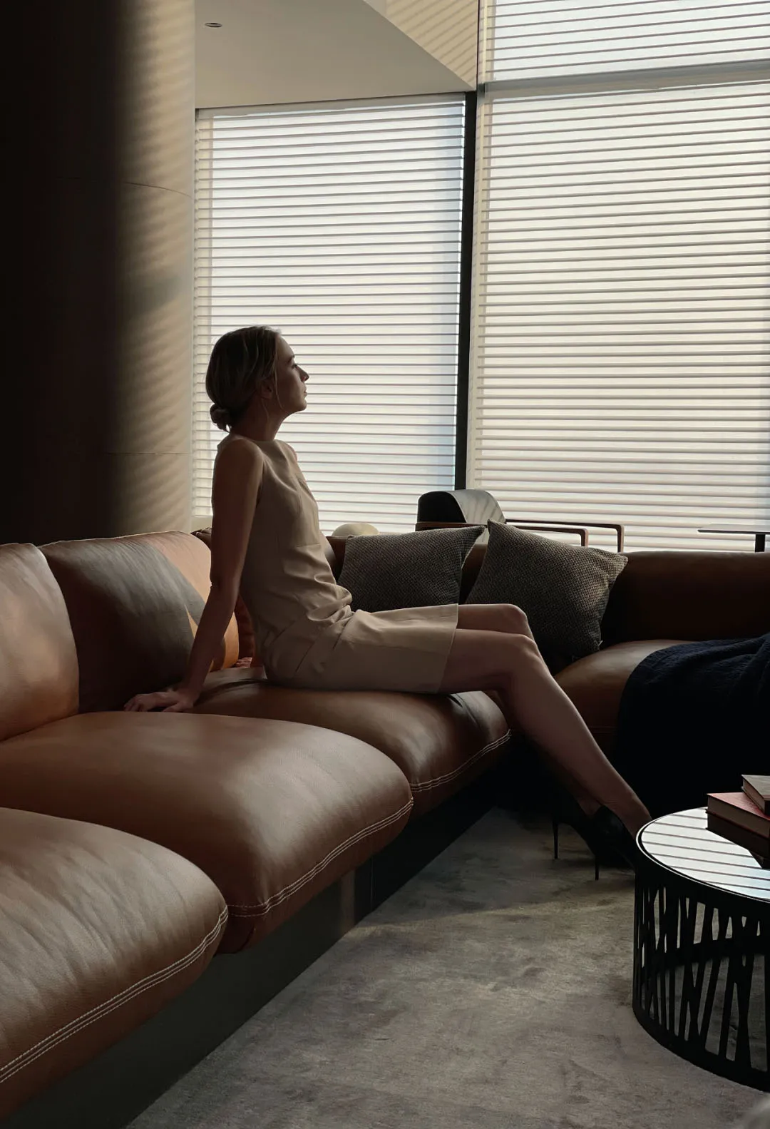 A woman in a sleeveless beige dress sitting on a modern leather sofa in a contemporary relaxation space with blinds.
