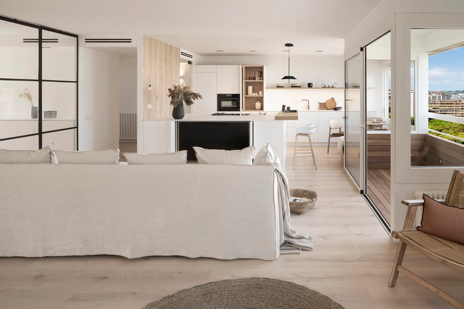 A modern living room with a white interior, featuring a large sofa, a kitchen island, and a balcony with a view of the city.