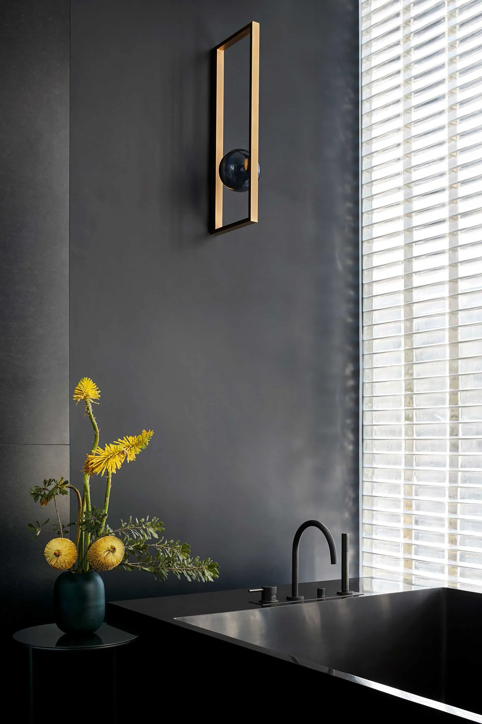 A sleek, modern bathroom featuring dark glass walls, a metal sink fixture, and a vase of yellow flowers on a side table.