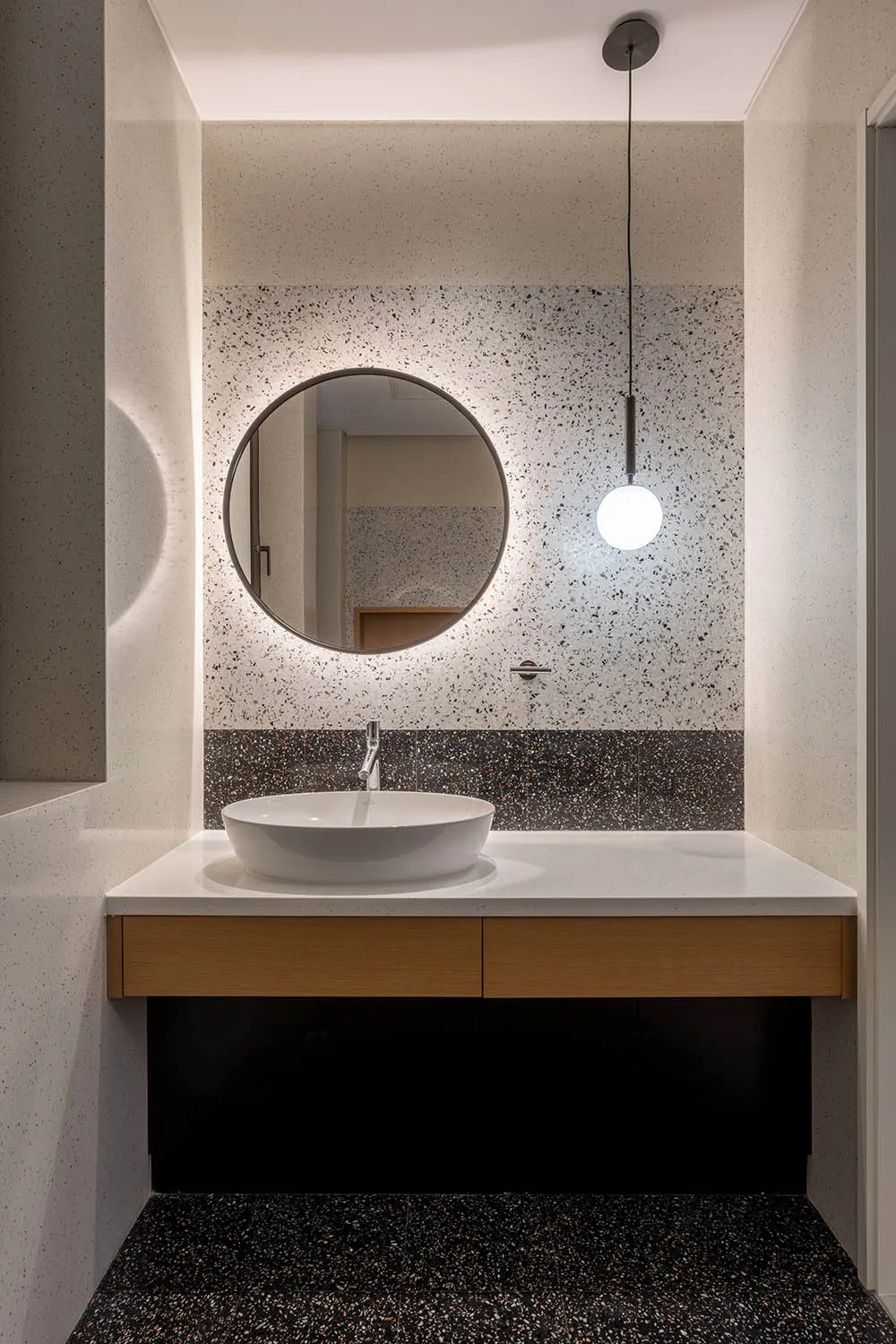 A modern bathroom featuring a terrazzo countertop and floor, with a circular mirror and pendant light.