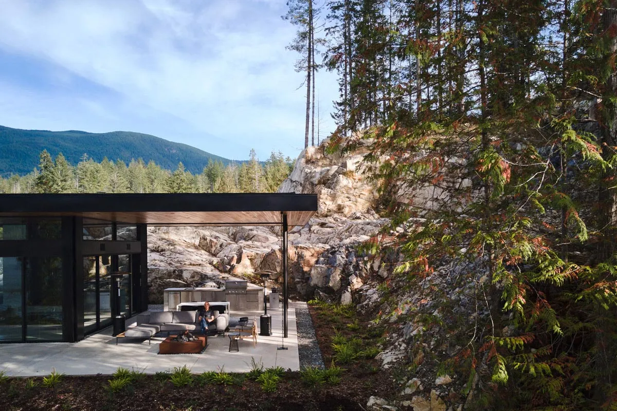 A modern house with concrete and glass features a spacious outdoor patio with a fire pit and lounge area.