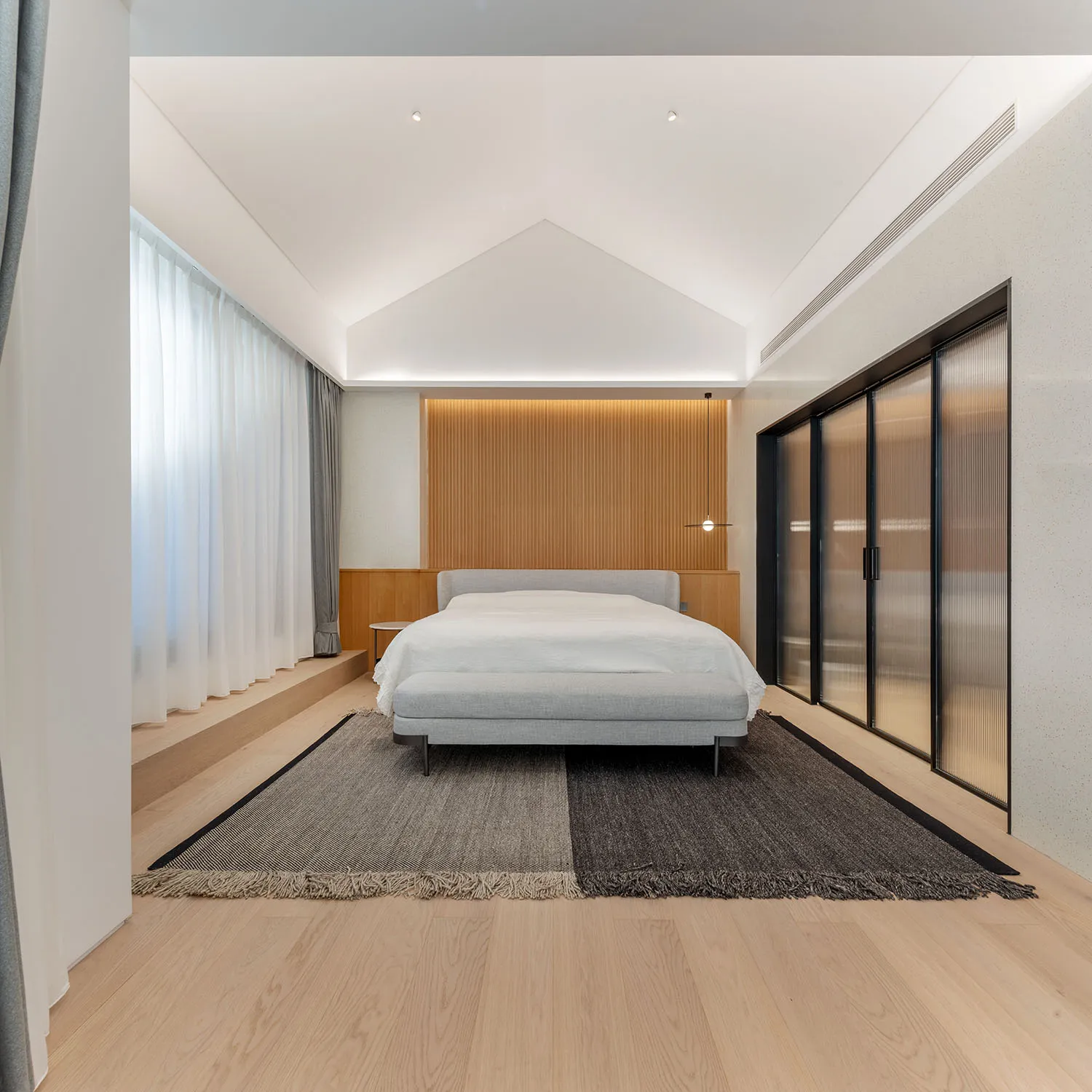 Modern minimalist bedroom with wood flooring, a bed with a minimalist grey headboard, and a rug with a clear geometric patter