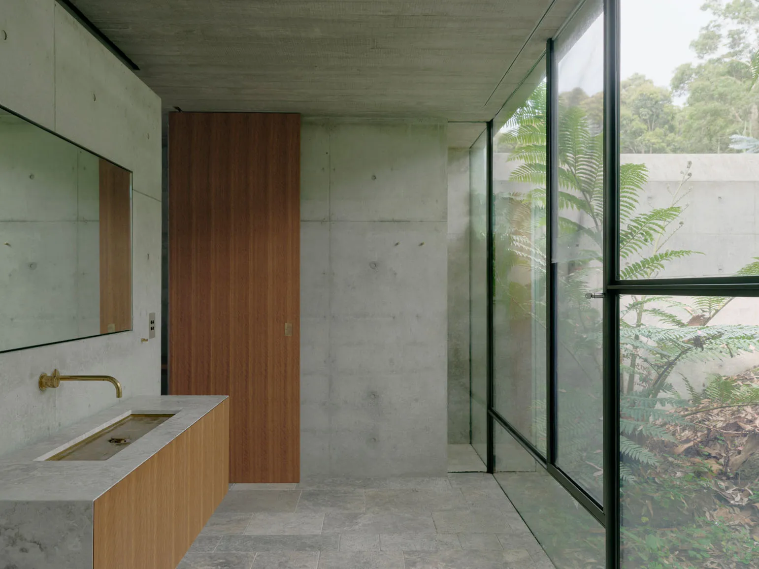 A modern bathroom with concrete walls, a wooden door, and large windows showing greenery outside.