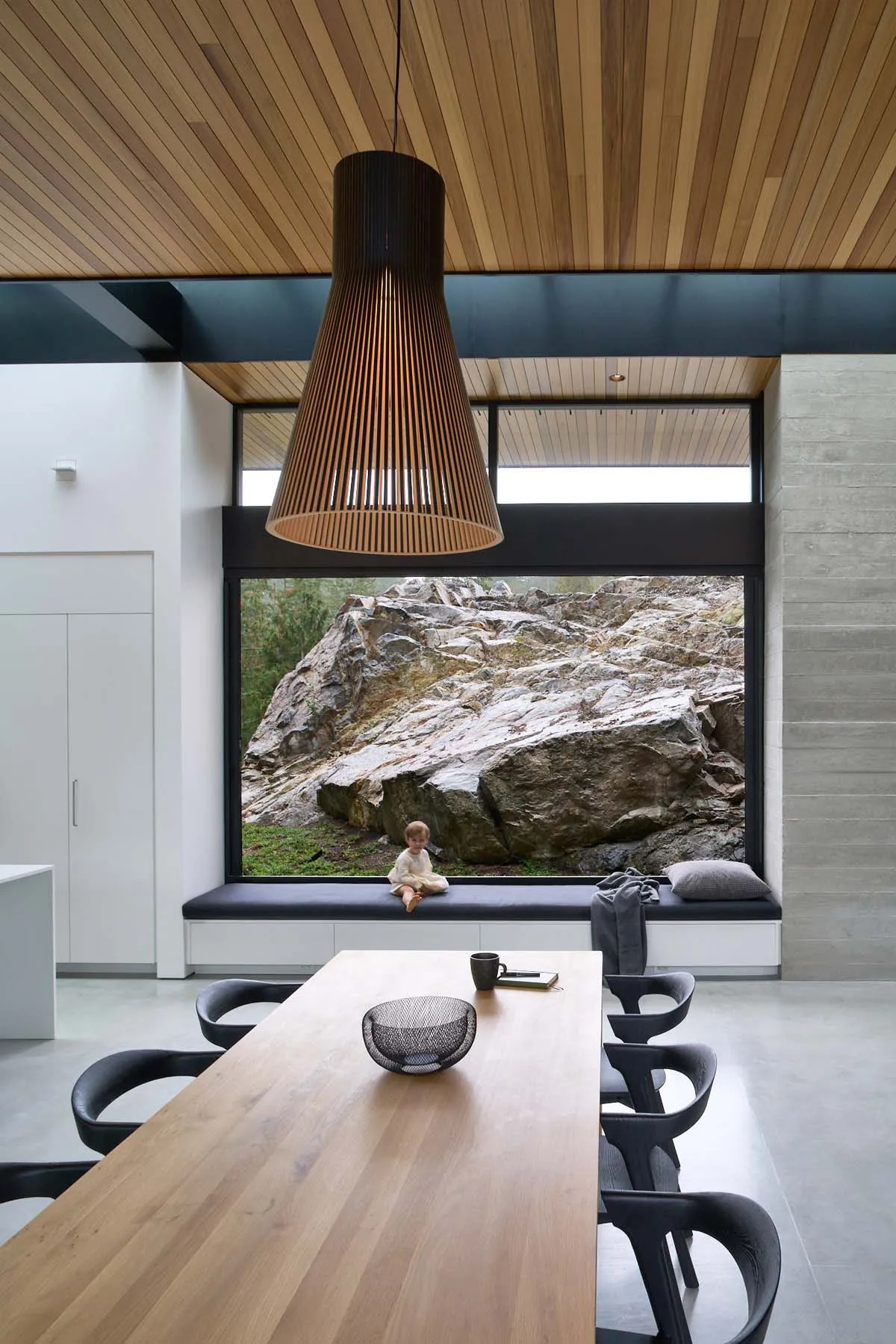 A modern dining room with a wooden table and seating area, featuring a large window with a view of rocks and nature outside.