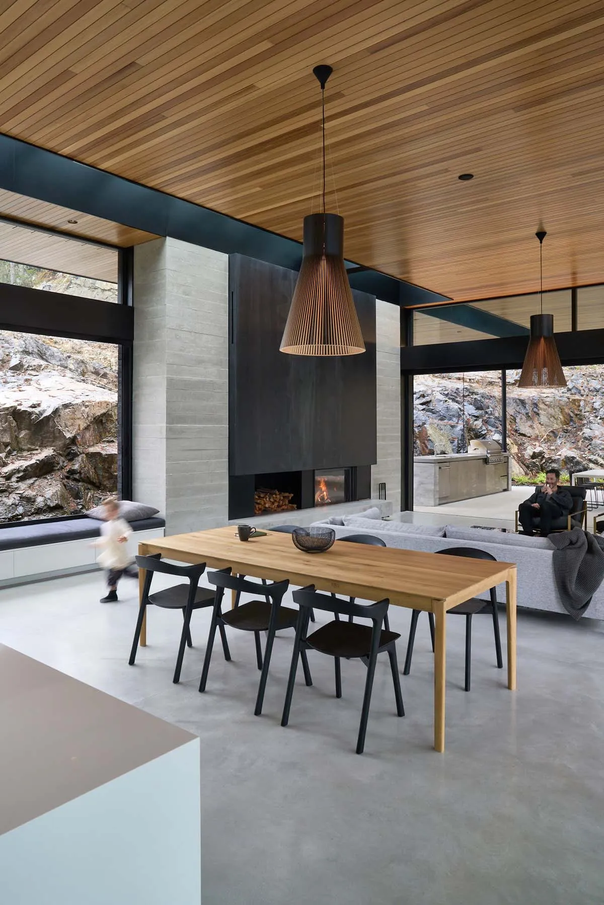 A modern, open-plan living and dining room featuring a wood dining table, concrete fireplace, and contemporary pendant lights