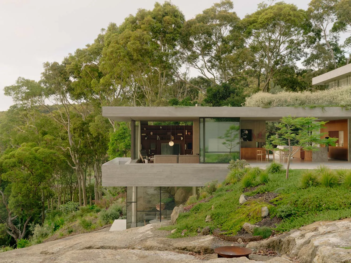 A modern minimalist house with concrete and glass features set in a natural, forested landscape.