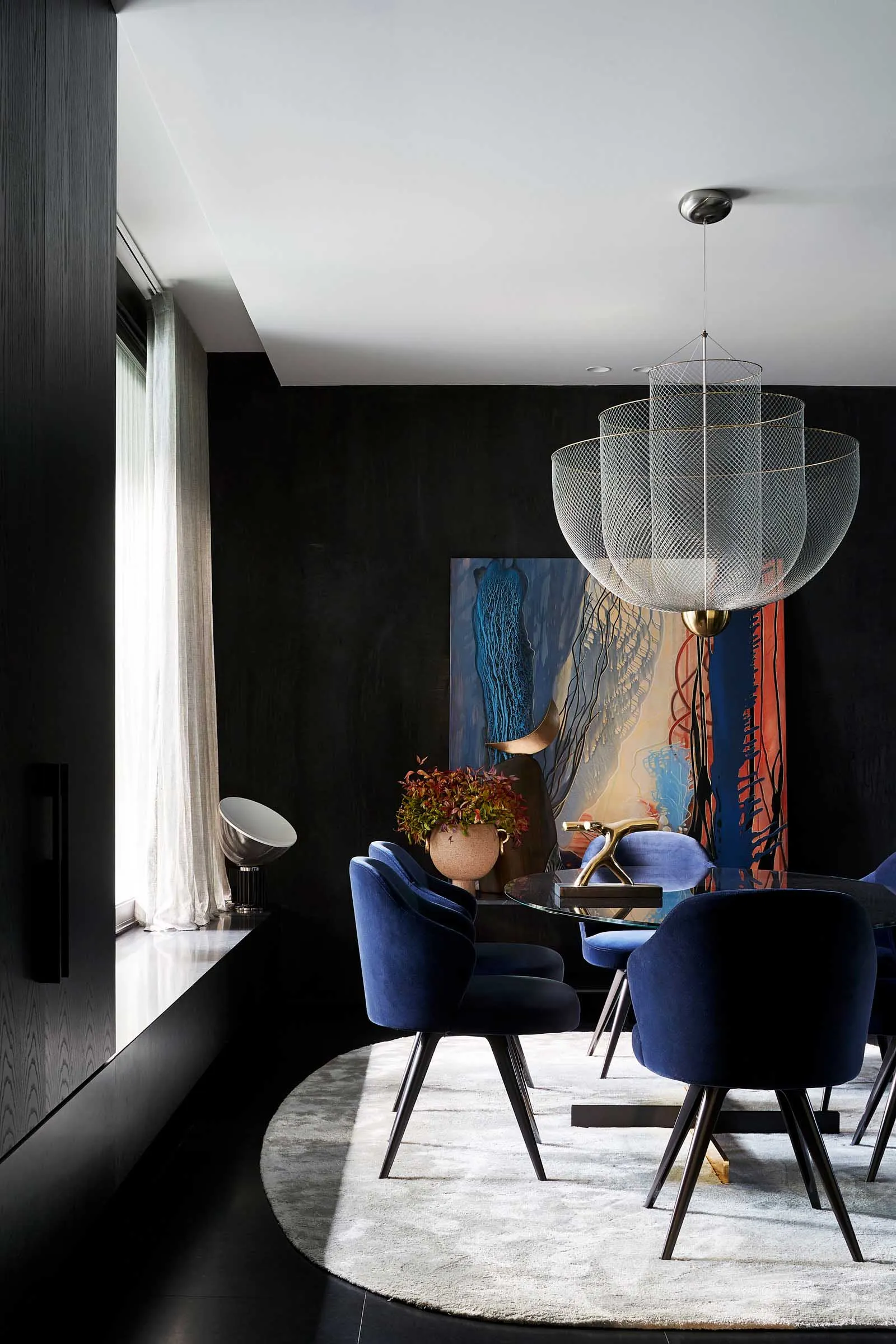 A modern interior design featuring a black wall, glass table, blue velvet chairs, and a large abstract painting.