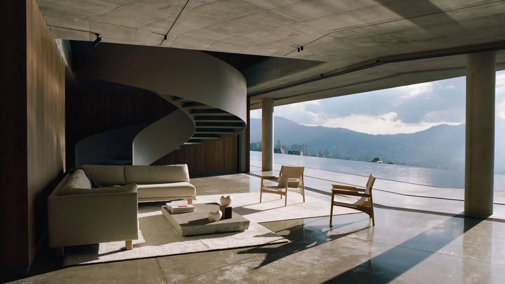 A modern living room featuring concrete walls and a glass spiral staircase overlooking mountains.
