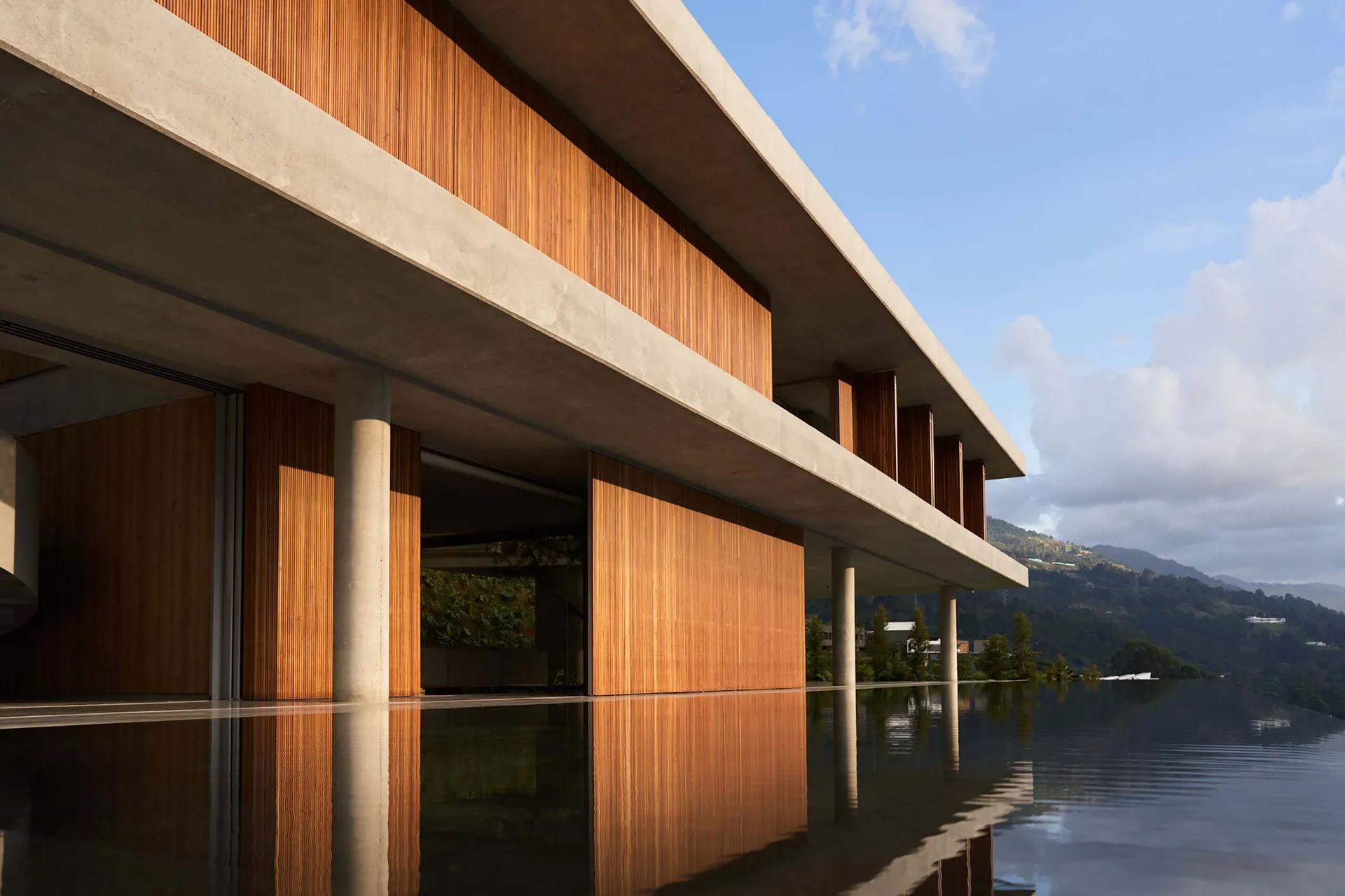 Modern architectural building with concrete structure and wooden accents reflected in a water body, set against a mountainous