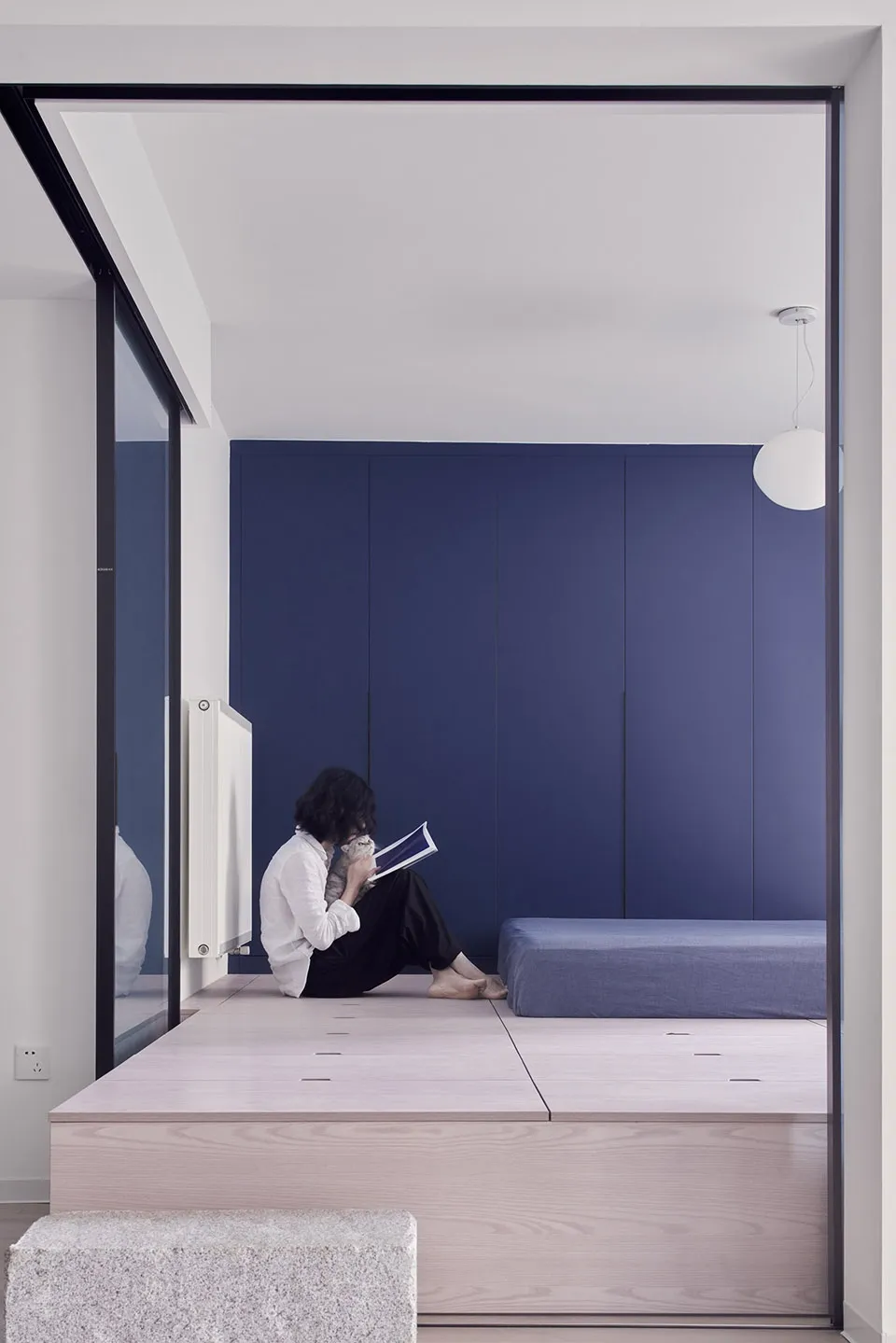 A minimalist living space featuring a wooden platform and a person sitting on it reading a book.