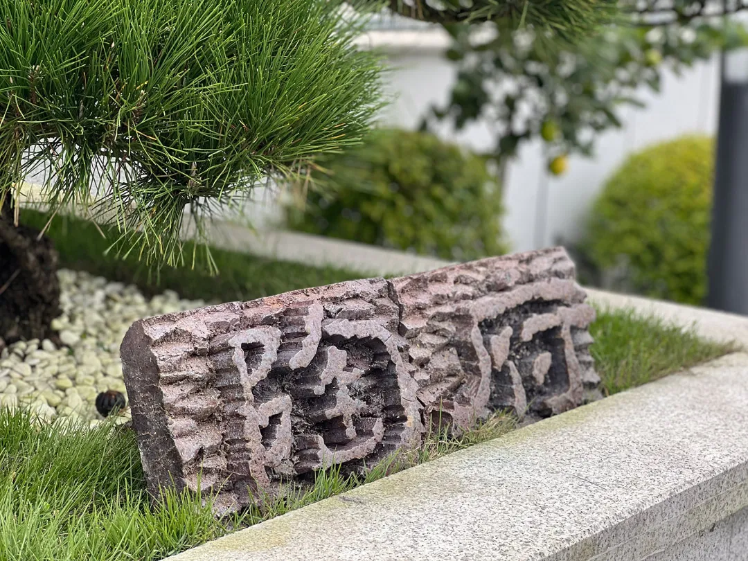A textured rock sculpture made of concrete with carved Chinese characters on a concrete ledge surrounded by greenery and gras