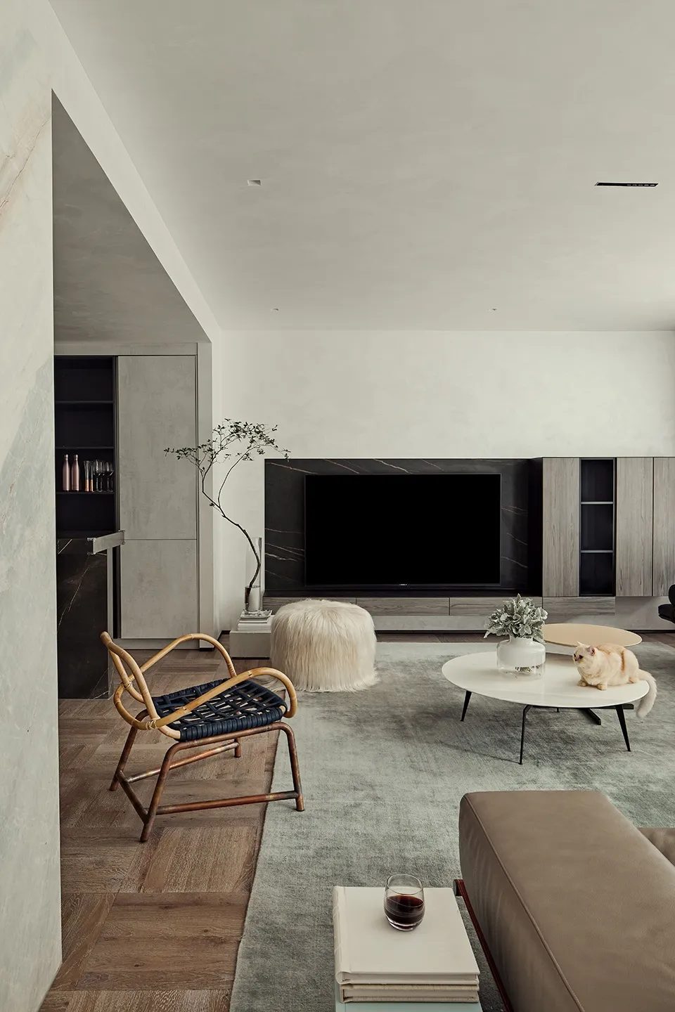 A minimalist living room with woven armchair, wooden sofa, fur pouf, and coffee table on a grey rug.