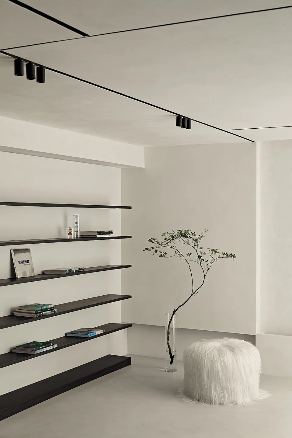 A minimalist living space featuring a concrete floor, floating shelves, and a white fur pouf with a potted plant.