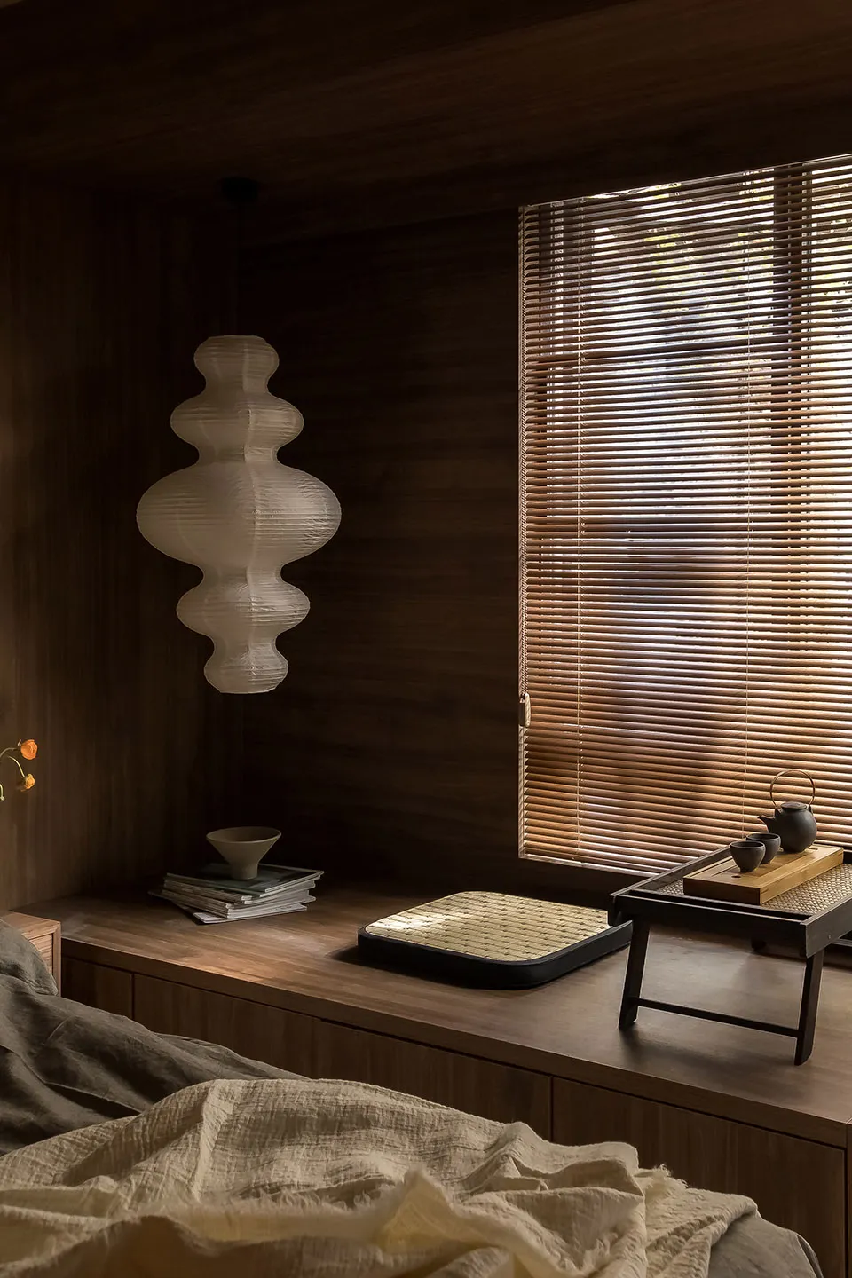 A minimalist bedroom featuring wooden accents, a window with blinds, and a lamp with a geometric design.