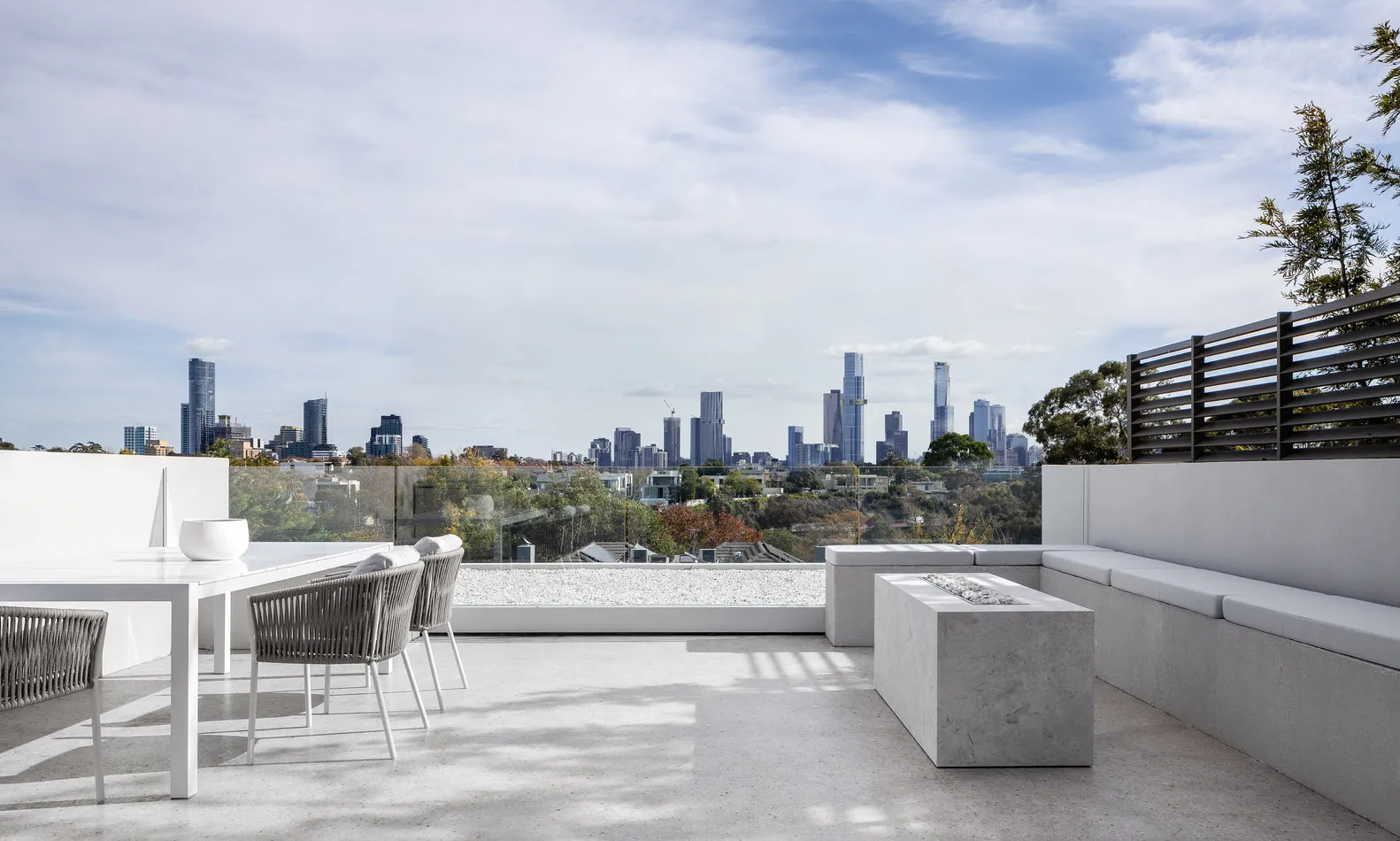 Modern outdoor terrace featuring grey concrete seating and a glass railing overlooking a city skyline.