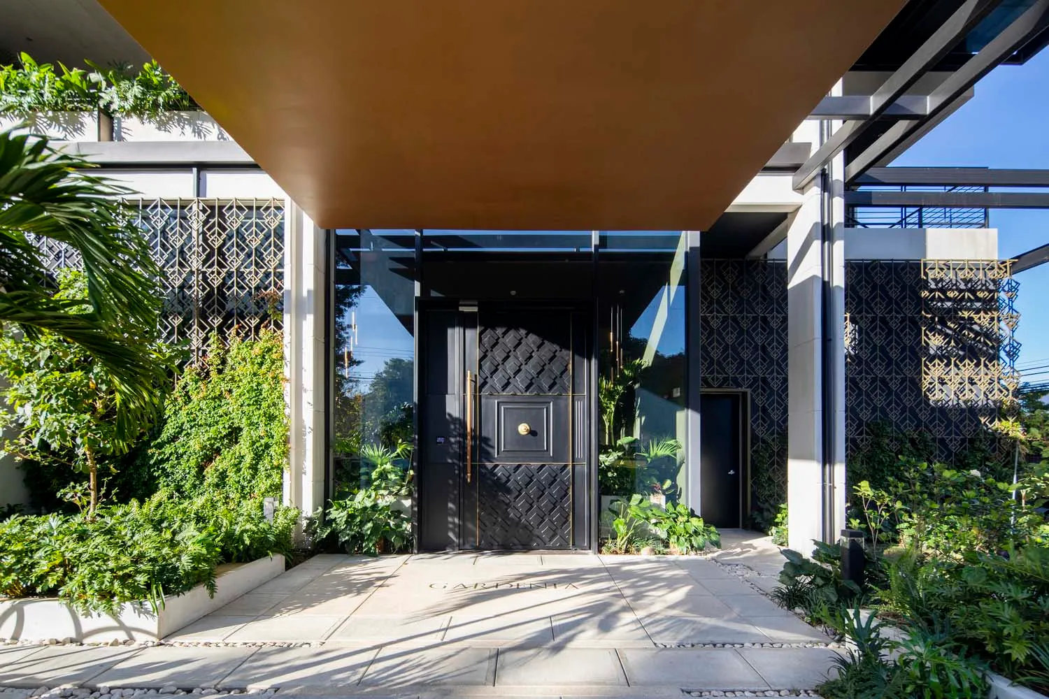A modern entrance featuring glass and metal materials, with greenery and a geometric pattern on the walls.