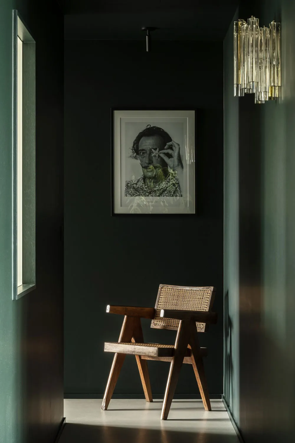 A minimalist interior design featuring a wooden chair and a black and white portrait hanging on a dark green wall.