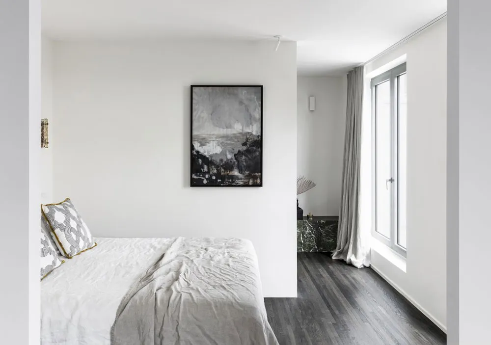 A minimalist bedroom with a white bed, gray walls, and a large window.