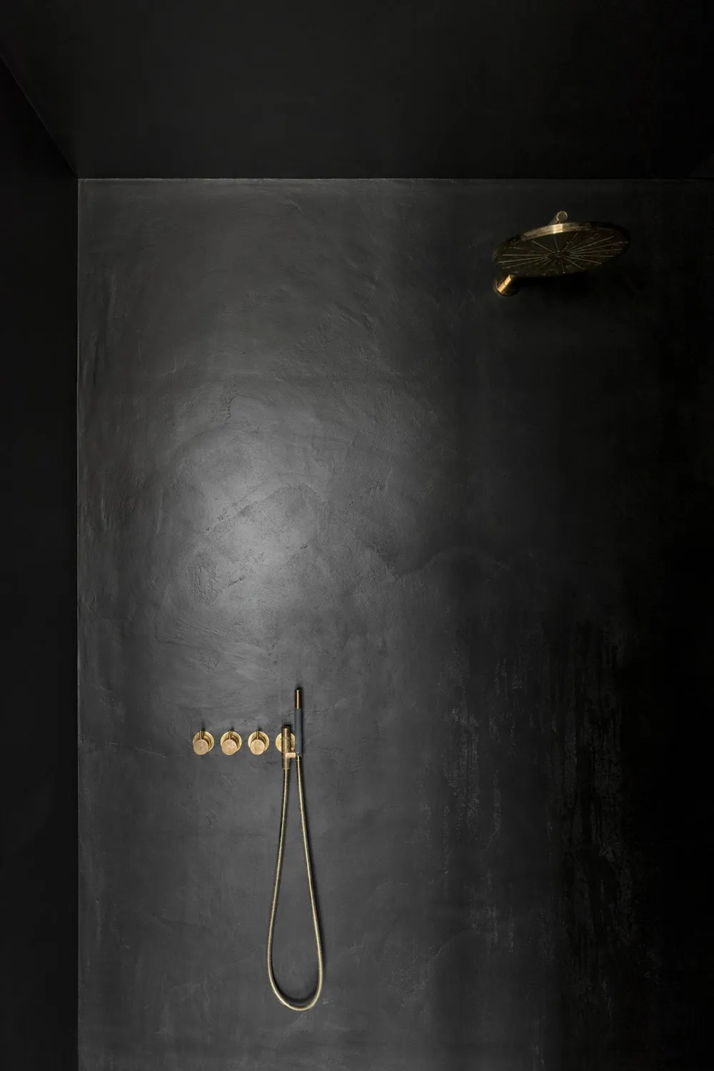 A modern bathroom with black concrete walls and brass fixtures, including a rain shower head, handheld showerhead, and push b