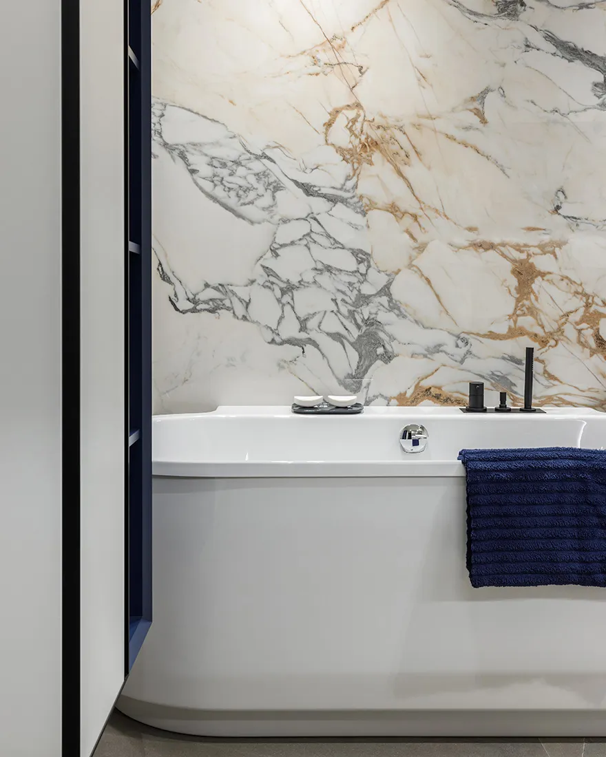 Modern minimalist bathroom featuring white marble walls and a white bathtub with blue towels.
