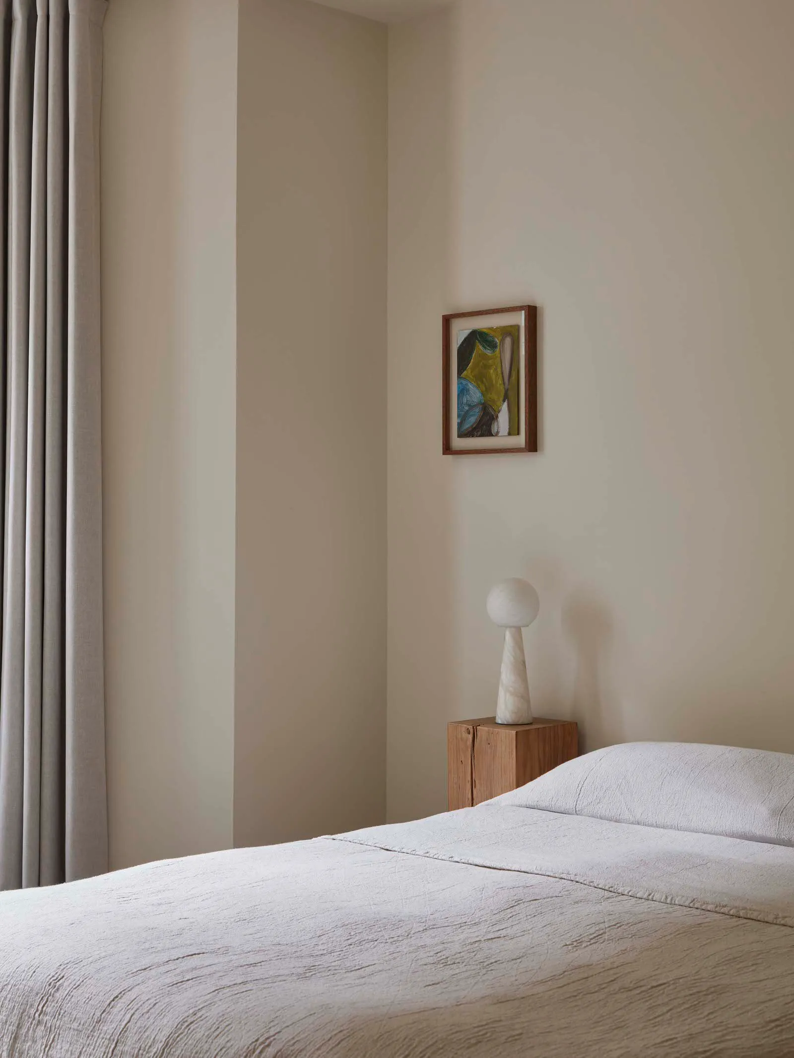 A minimalist bedroom features grey drapery, a wooden bed frame, and a marble lamp on a wooden nightstand.