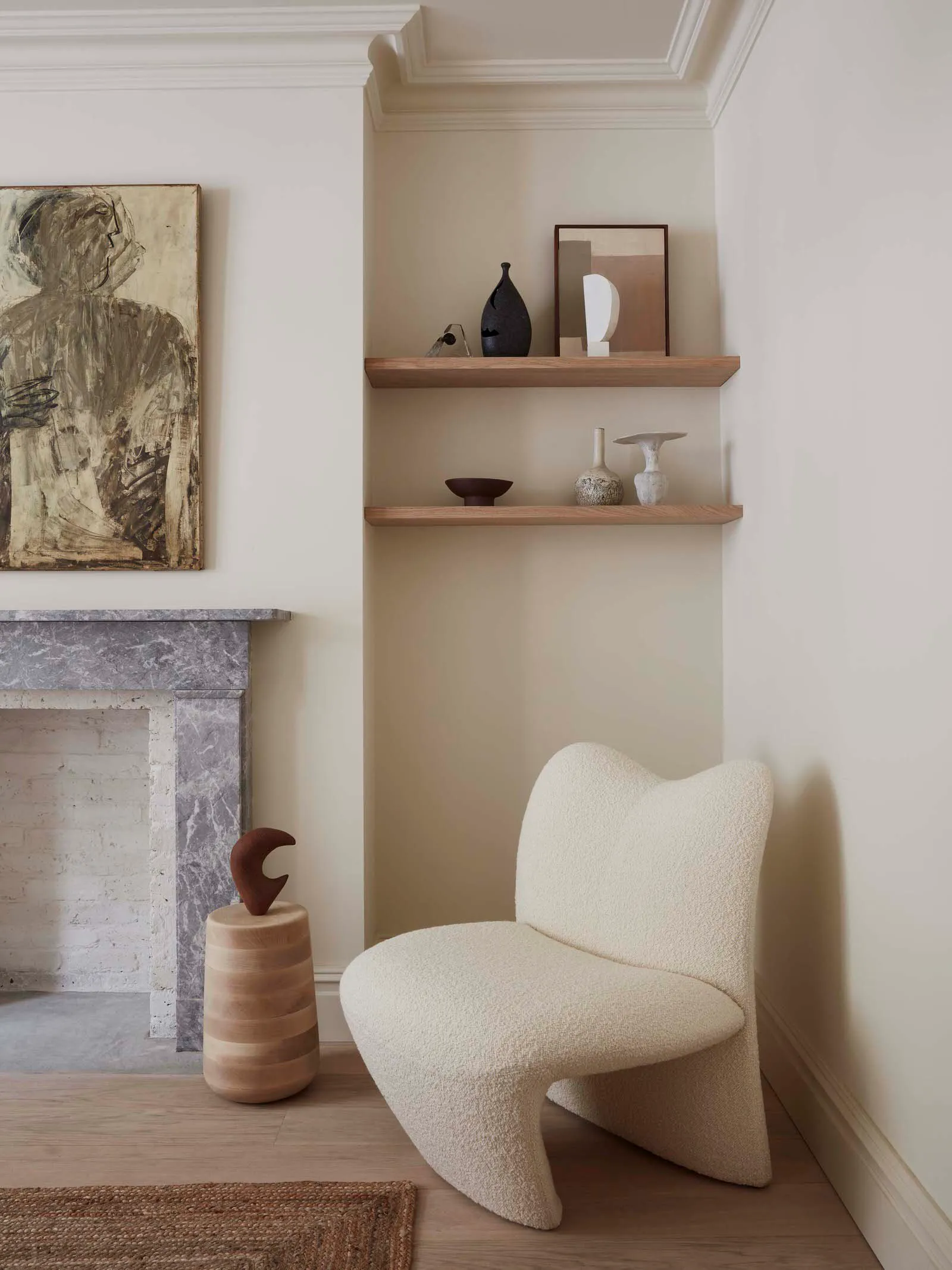 A cozy corner of a minimalist residential interior featuring a white textured lounge chair, a marble fireplace, wood shelves,