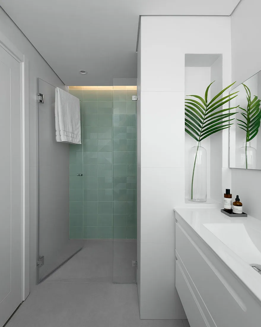 Minimalist bathroom with green glass tile shower, white tiles, and concrete floor, featuring a floating vanity and a leafy pl