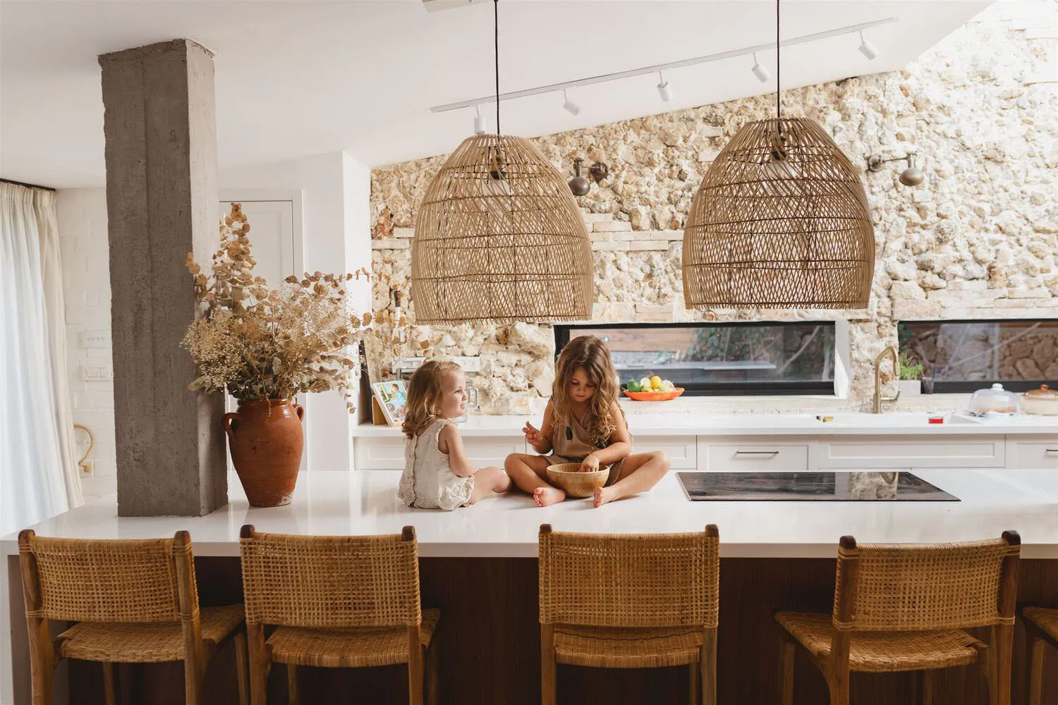 Two children sitting on a kitchen island with wooden chairs, decorated with dried flowers in a vase and large woven pendant l