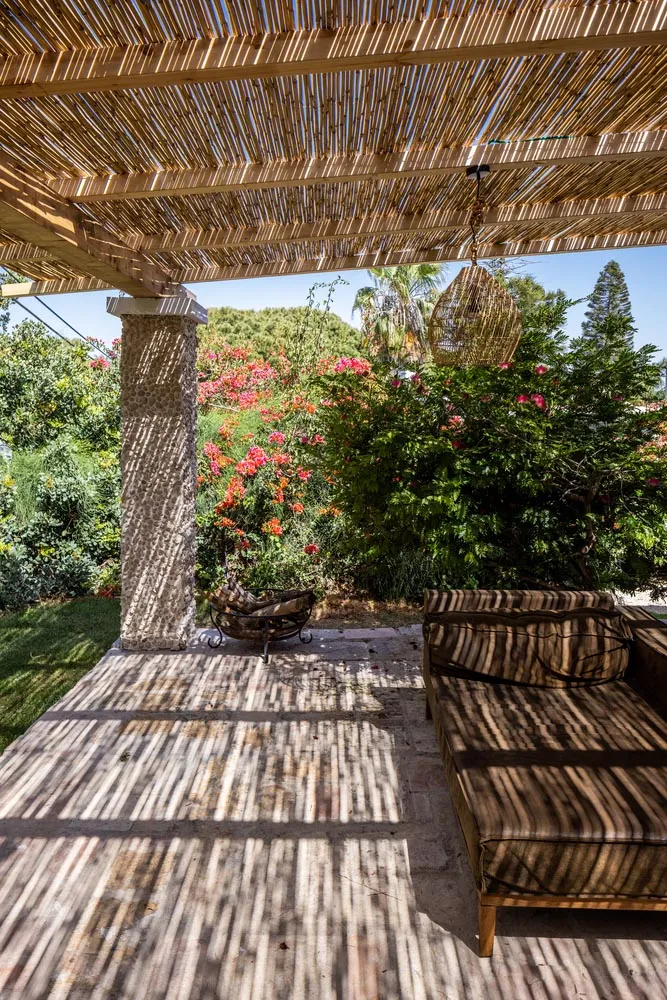 Outdoor wooden pergola with wicker furniture and cement bench surrounded by lush greenery and blooming flowers.