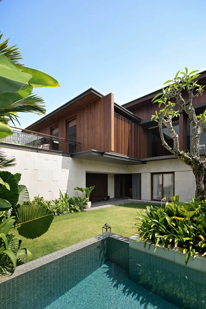A modern residential pool house with a concrete, wood, and tile exterior, featuring lush landscaping and a swimming pool surr