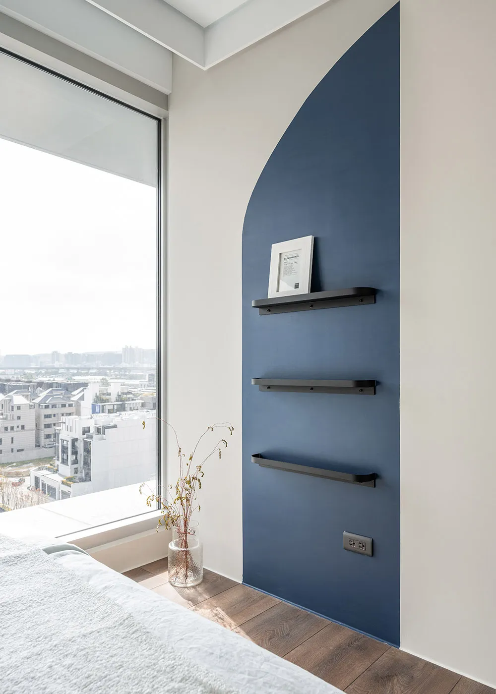 A modern bedroom featuring a blue accent wall with sleek, black metal shelves and a large window overlooking a cityscape.