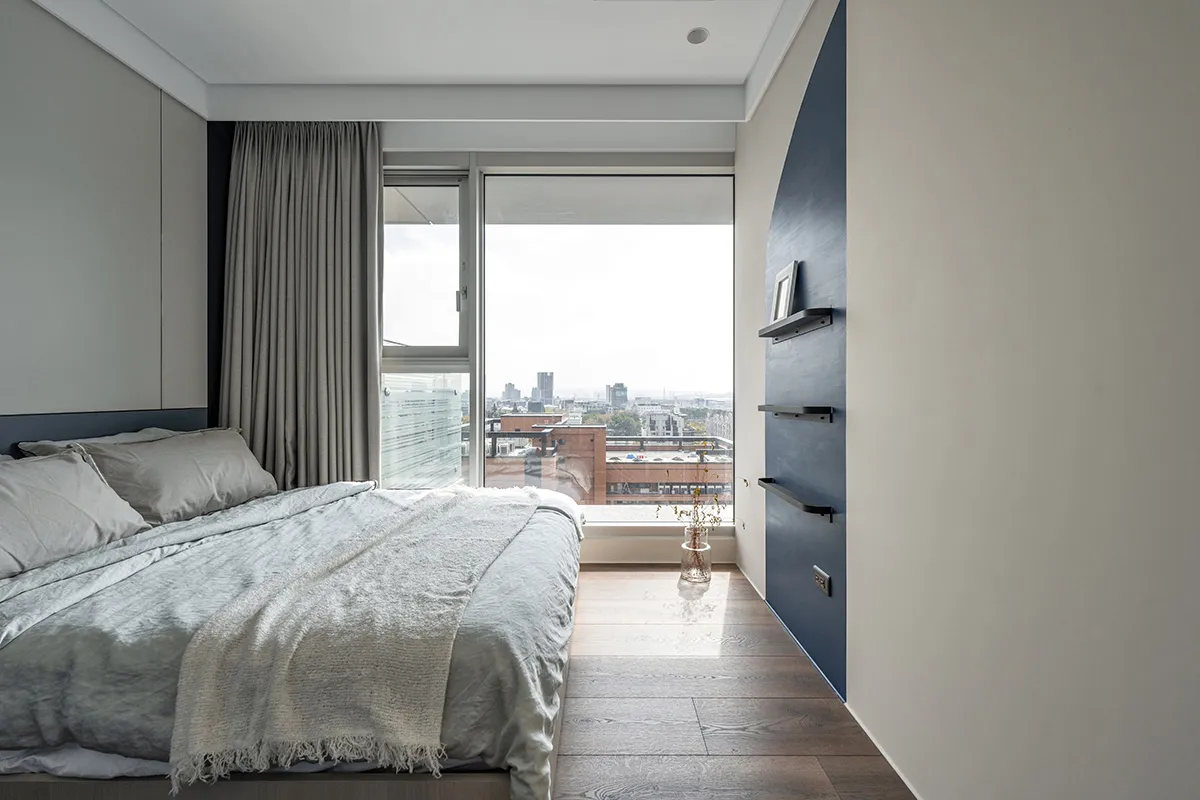 A modern, minimalist bedroom with timber flooring, a large window overlooking a cityscape, and simple furniture.