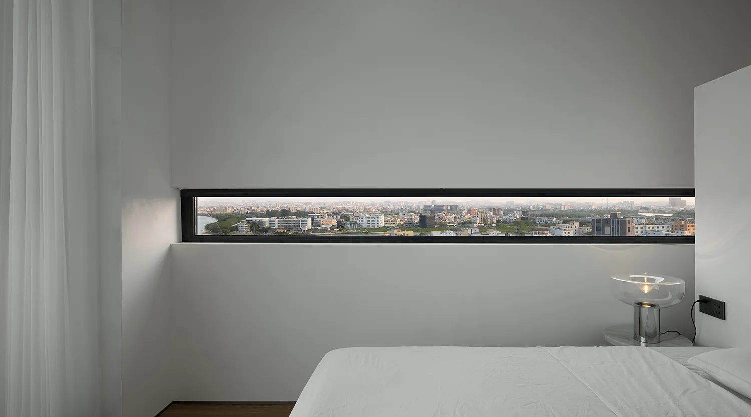 Modern minimalist bedroom featuring white walls and a city view through a horizontal window.