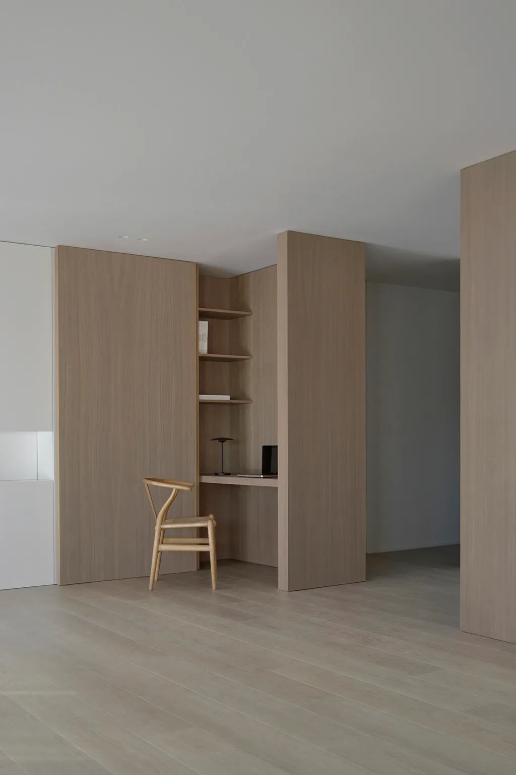 A minimalist office space featuring a desk, chair, and shelves, with a focus on open space and natural light.