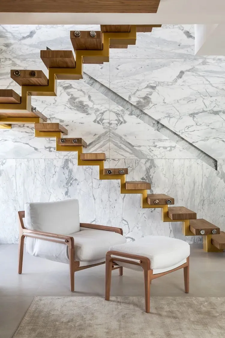 A modern interior space featuring a marble staircase with wooden steps and a white cushioned armchair and ottoman.