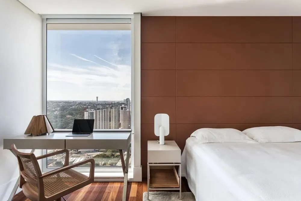 Modern bedroom with a large window overlooking a cityscape, featuring a white bed, white desk, and brown wooden flooring.