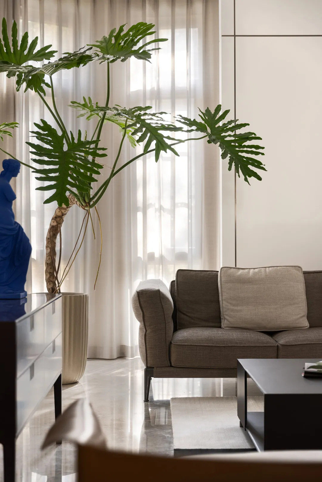Modern living room featuring a beige fabric sofa, white curtains, and metal coffee table with plants and a statue.