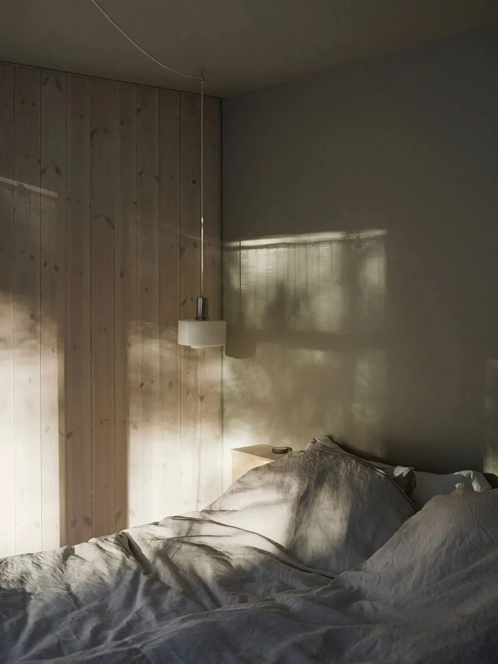 A minimalist bedroom with wooden walls and fabric bedding, featuring natural light and a hanging lamp.