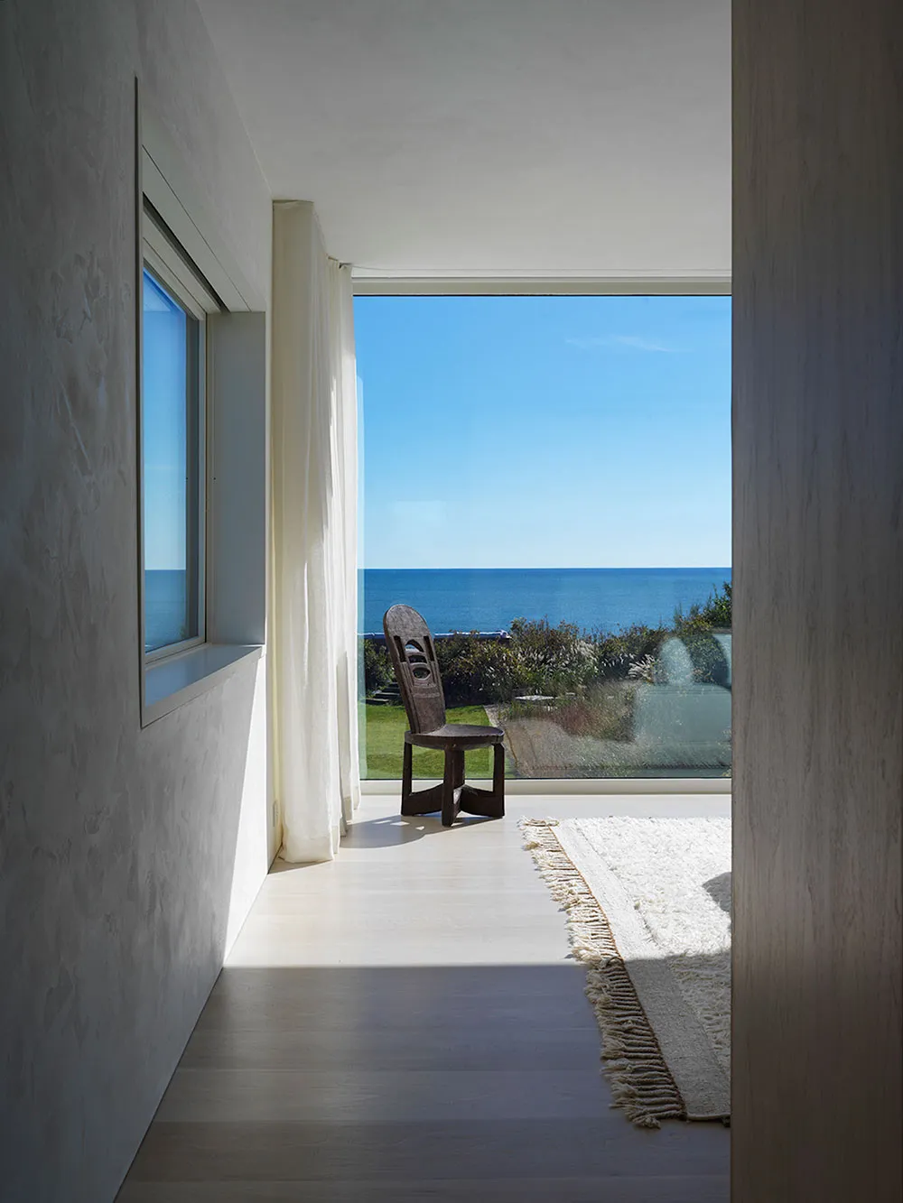 A minimalist residential space with a beach view featuring a wooden chair, glass window, and white fabric rug.