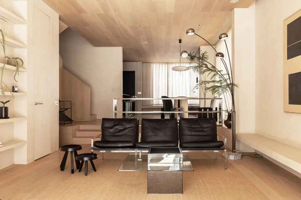 Modern minimalist living room featuring wood-paneled walls, black leather sofas, glass coffee table, and potted plants.
