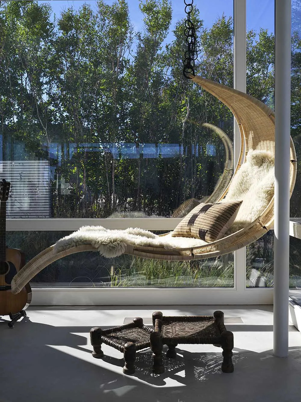 A modern indoor swing with a wicker frame and plush cushions hanging from a ceiling hook, surrounded by large windows with a