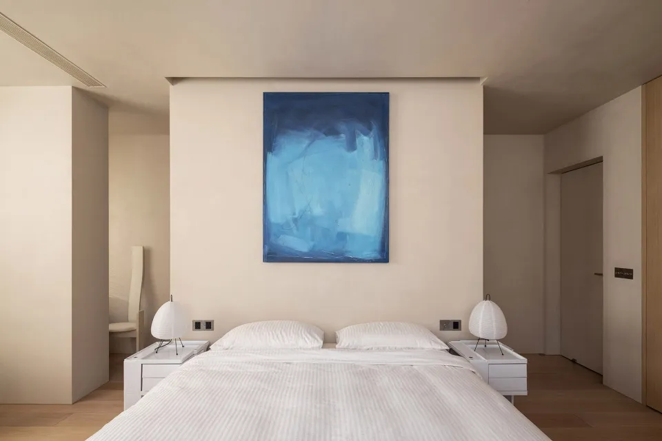 Modern minimalist bedroom with a blue abstract painting above the bed, white bedding, and two cylindrical lamps on side table